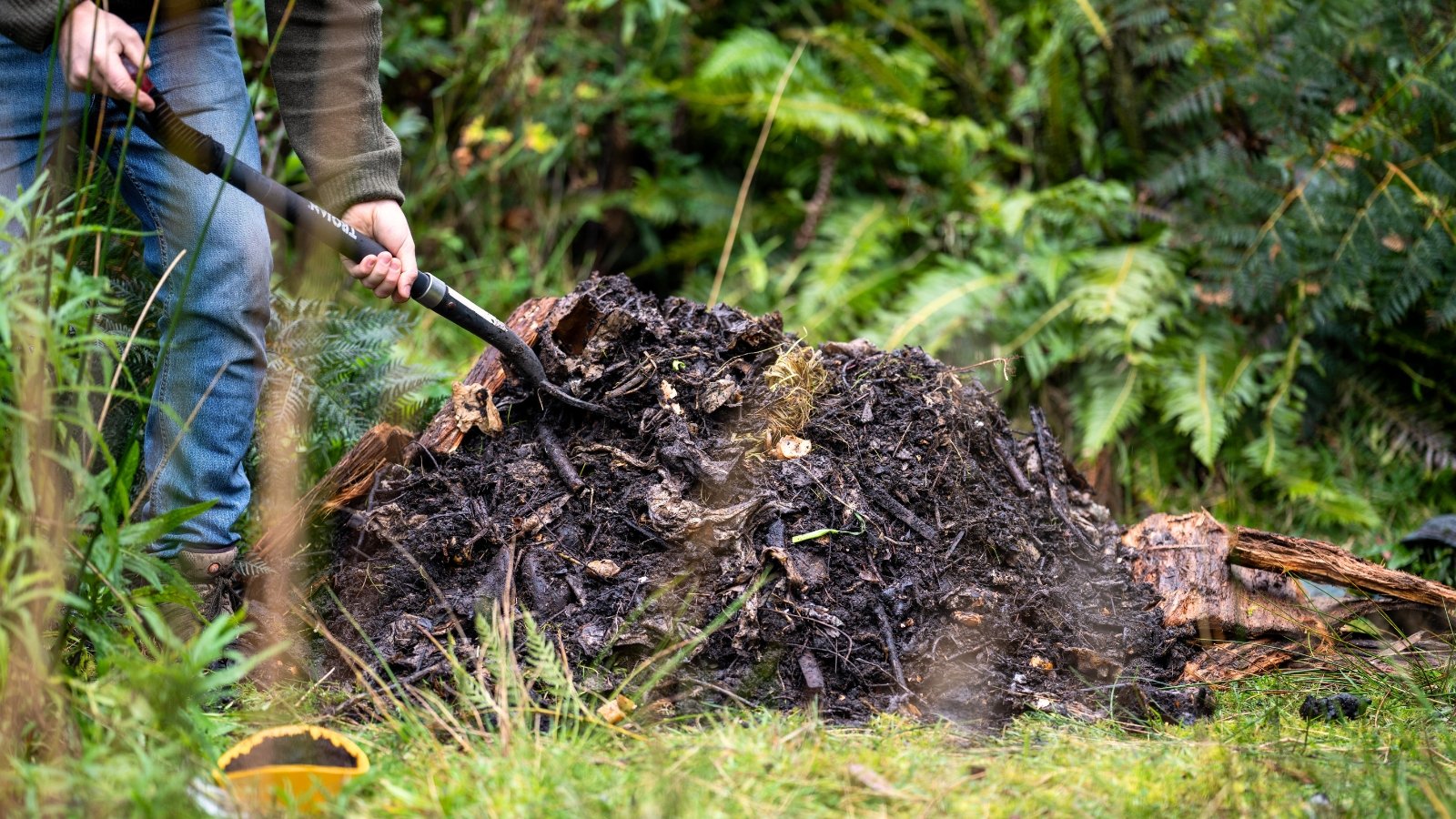 Close-up of a gardener wearing gloves, using a pitchfork to turn over a dark pile in the garden, revealing a mix of decomposing organic matter.